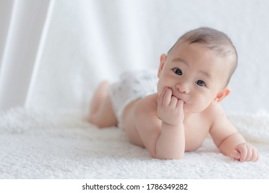 Asian Baby Boy Lying On His Tummy On A Piece Of White Fluffy Cloth Chewing His Hands And Smiling At The Camera