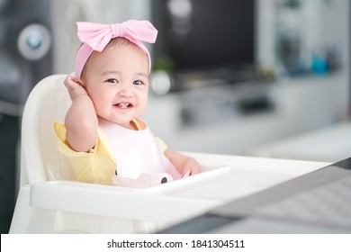 Asian Baby Girl In A Pink Bib And Hair Bow Sitting On A White High Chair Is Happy And Enjoy During Having A Breakfast At Home