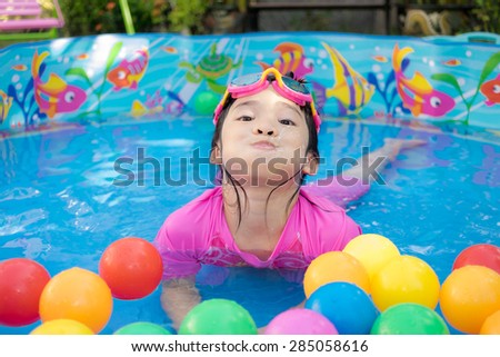 An asian baby girl in pink suit playing water and colorful balls in blue kiddie pool. She's smiling, happy and having fun