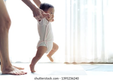 Asian baby taking first steps walk forward on the soft mat. Happy little baby learning to walk with mother help at home. Mother teaching how to walk gently. Baby growth and development concept.