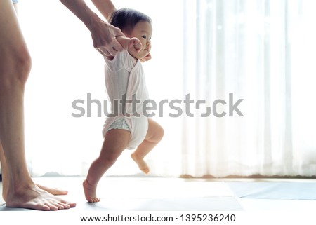 Asian baby taking first steps walk forward on the soft mat. Happy little baby learning to walk with mother help at home. Mother teaching how to walk gently. Baby growth and development concept.