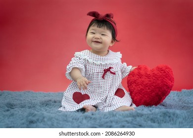 Asian Baby Valentine With Red Heart Smiling And Sitting On Red Background. Cute 6 Months Baby Sitting With Copy Space Use As Concept Of Valentine, Love, Baby Or Kid Department In Hospital.