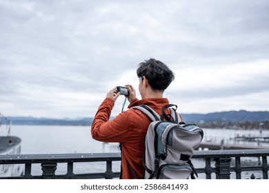 Asian backpacker use camera taking a picture of beautiful landscape. Young male tourist traveler travel alone on street in city, using cellphone record vlog on holiday vacation trip in Switzerland.