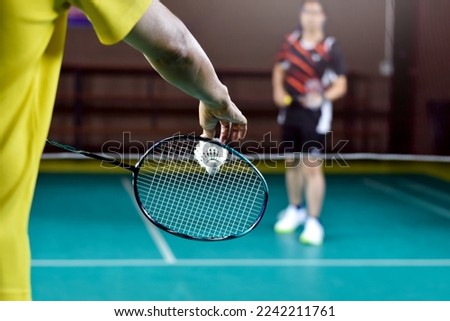 Asian badminton player holds racket and white cream shuttlecock in front of the net before serving it to another side of the court.