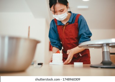 Asian Baker In Her Bakery Wearing A Mask While Picking Up A Mini Cake.