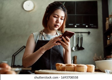 Asian Baker Woman Taking Photo Of Fresh Cupcake On Wooden Plate In The Kitchen With Smartphone For Social Media.