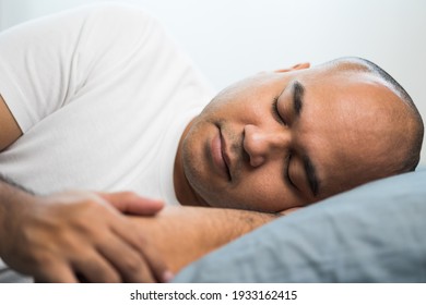 An Asian bald man around the age of 30 in a white T-shirt is sleeping on his side on a blue mattress and pillows. He slept at home until morning.
