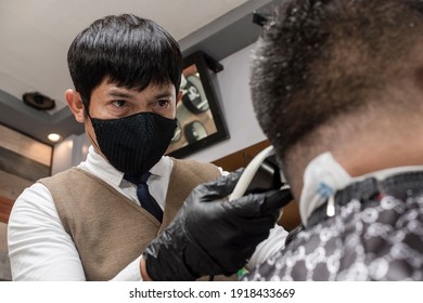 An Asian Barber Wearing A Face Mask Trims And Fades The Back Of A Client's Head With A Hair Clipper.