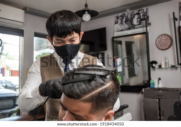 An asian barber wearing a face mask give a
regular client a haircut at his
salon.