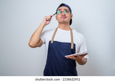 Asian Barista Man In Blue Apron Taking Order, Writing On Menu Book List, Thinking With Pen On Head. Isolated Image On White Background