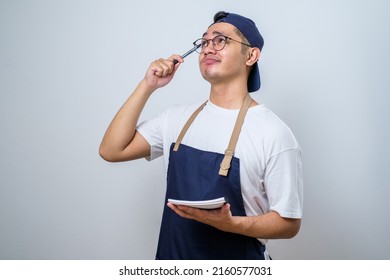 Asian barista man in blue apron taking order, writing on menu book list, thinking with pen on head. Isolated image on white background - Powered by Shutterstock