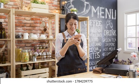 Asian barista woman standing success sme with cute smiling at city modern cafe and holding a smart phone in good day so happy. cheerful attractive girl waitress laughing and looking at cellphone.