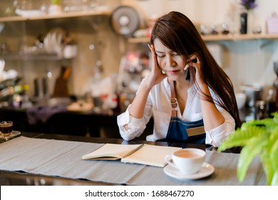 An Asian barista woman is stressed because of economic conditions and COVID-19 or Coronavirus has caused her sales in the coffee shop to drop dramatically, the impact of a small self-employed business