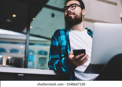 Asian Bearded Hipster Guy Holding Mobile Phone Making Money Transaction Doing Shopping Online In Market,man Millenial In Eyewear Looking Away Using Laptop Computer And Smartphone For Business Outdoors