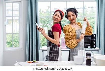 Asian Beautiful Adorable Two Women Or Friends Wearing Aprons, Holding Bakery Equipments, Listening Music, Smiling, Cooking And Dancing With Fun And Happiness In Kitchen At Comfortable Home On Weekend