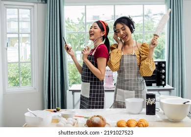 Asian Beautiful Adorable Two Women Or Friends Wearing Aprons, Holding Bakery Equipments, Listening Music, Smiling, Cooking And Dancing With Fun And Happiness In Kitchen At Comfortable Home On Weekend