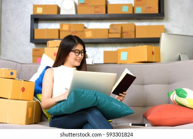 Asian Beautiful Charming Girl Entrepreneur Checking Order On Her Laptop And Writing To Memorandum Book. Business And Technology Concept.