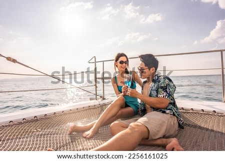 Asian beautiful couple drinking champagne while having party in yacht. Attractive man and woman hanging out, celebrating anniversary honeymoon trip while catamaran boat sailing during summer sunset.
