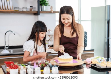 Asian beautiful female baker pastry chef mother wears apron standing smiling helping teaching little girl daughter decorating cake with whipping cream making homemade bakery in decorated home kitchen.