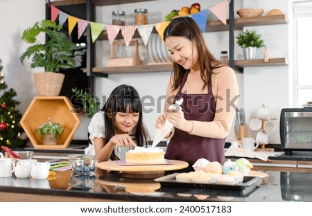Asian beautiful female baker pastry chef mother wears apron standing smiling helping teaching little girl daughter decorating cake with whipping cream making homemade bakery in decorated home kitchen.