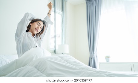 Asian beautiful girl in pajamas wake up in the morning with happiness. Attractive young woman smiling, feel happy and relax then stretching body after getting up from sleep on bed in bedroom at home.