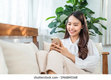 Asian beautiful girl with smile face sit on sofa in living room, spend leisure time in house. Young attractive woman feeling happy and relax stay at home texting chat message on smartphone with friend