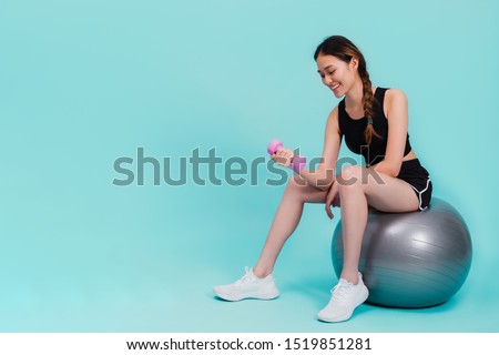 Asian beautiful happy woman holding dumbell and sitting on fit ball after exercise isolated on blue color background.Concept of slim and healthy girl workout.