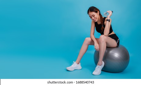 Asian Beautiful Happy Woman Wearing Smart Watch Holding Water Bottle And Sitting On Fit Ball After Exercise Isolated On Blue Color Background.Concept Of Slim And Healthy Girl Workout.