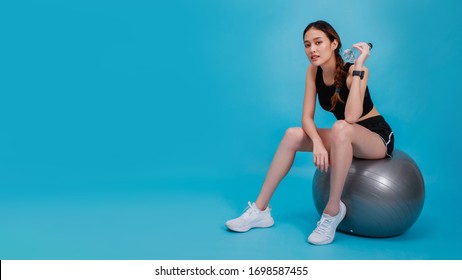 Asian Beautiful Happy Woman Wearing Smart Watch Holding Water Bottle And Sitting On Fit Ball After Exercise Isolated On Blue Color Background.Concept Of Slim And Healthy Girl Workout.