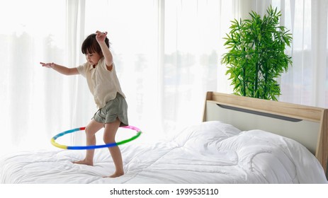 Asian beautiful little playful girl fail when try standing learning playing colorful hula hoop alone on white clean sheet bed in bedroom with green tree pot and clear curtain in background.
