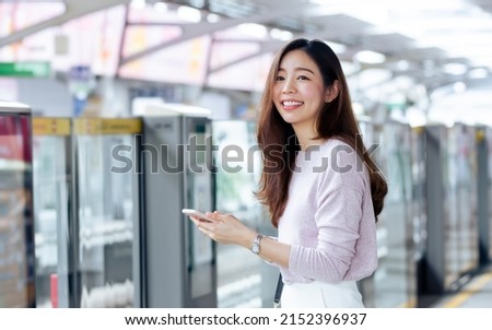 Asian beautiful long hair woman smiling with happiness and refresh, wearing casual business shirt, holding mobile phone, standing on platform, waiting for sky train transportation in morning day