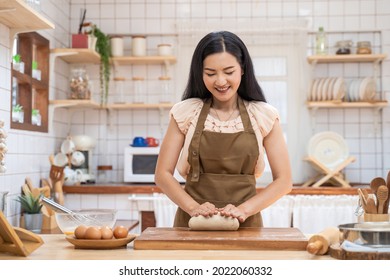 Asian beautiful mature woman stay home, spend time in kitchen cooking. Young attractive carring female wear apron and smile, kneaded yeast dough with hand to baking bakery on table for dinner in house