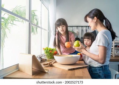 Asian Beautiful Mother Cooking Vegetable Salad With Two Young Daughter. Happy Family, Attractive Caring Parent Mom Enjoy Preparing Healthy Foods For Little Girl Child Sibling Sister In Kitchen At Home