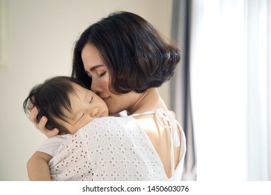 Asian Beautiful Mother Hugging Sleeping Baby In Her Arms And Kissing The Kid Gently. The Mom Closing Her Eyes While Holding Her Baby Head To Rest On Shoulder. Touch Of Love And Family Relationship.