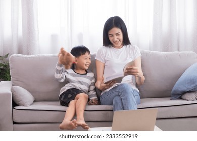 Asian beautiful mother and son read favorite book together on weekends, young woman and adorable boy lying down on sofa reading a comic book in living room, Enjoy listening to stories from mom.