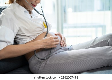 Asian Beautiful Pregnant Woman Sit On Sofa And Listen Baby's Heartbeat. Attractive Young Pregnancy Mother Feeling Happy And Excited While Using Stethoscope Listen To Newborn Child Vital Signs In House