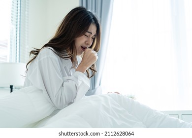 Asian beautiful sick girl in pajamas getting up from sleep in bedroom. Attractive young woman feeling bad after wake up on bed and coughing in early morning at home. health care-lifestyles concept. - Shutterstock ID 2112628064