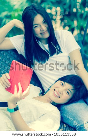 Asian Beautiful Woman Beauty Face is Lying and Holding Computer Tablet on Lap of Her Friend on the Swing in Garden. Idea Concept for Relaxation on Holiday or Vacation. Vertical Style.