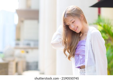 Asian Beautiful Woman With Bronze Long Hair In Purple Camisole And White Sleeve Stands On The Street As Background.