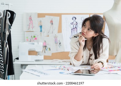 Asian Beautiful Woman Fashion Designer Or Dressmaker Thinking While Drawing Sketch Of New Collection Clothes On Paper, Working With Tablet On Desk At Workplace Studio