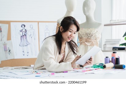 Asian Beautiful Woman Fashion Designer Or Dressmaker Drawing Sketch Of New Collection Clothes And Smiling While Working With Tablet On Desk At Workplace Studio