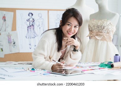 Asian Beautiful Woman Fashion Designer Or Dressmaker Drawing Sketch Of New Collection Clothes, Looking At Camera And Smiling While Working With Tablet On Desk At Workplace Studio