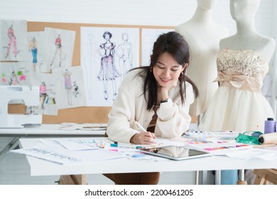 Asian Beautiful Woman Fashion Designer Or Dressmaker Drawing Sketch Of New Collection Clothes On Paper And Smiling While Working With Tablet On Desk At Workplace Studio