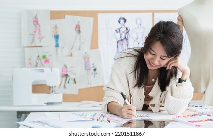 Asian Beautiful Woman Fashion Designer Or Dressmaker Drawing Sketch Of New Collection Clothes On Paper And Smiling While Working With Tablet On Desk At Workplace Studio