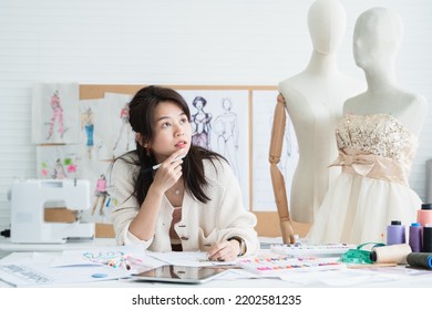 Asian Beautiful Woman Fashion Designer Or Dressmaker Thinking While Drawing Sketch Of New Collection Clothes On Paper, Working With Tablet On Desk At Workplace Studio