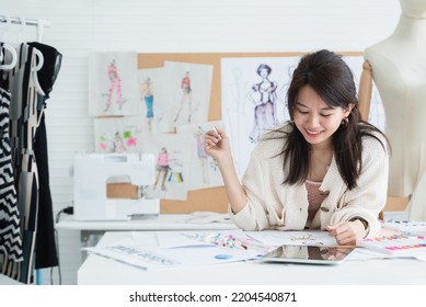 Asian Beautiful Woman Fashion Designer Or Dressmaker Drawing Sketch Of New Collection Clothes On Paper And Smiling While Working With Tablet On Desk At Workplace Studio