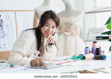 Asian Beautiful Woman Fashion Designer Or Dressmaker Drawing Sketch Of New Collection Clothes, Looking At Camera And Smiling While Working With Tablet On Desk At Workplace Studio