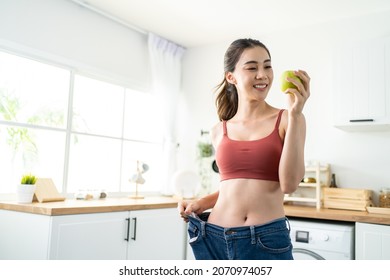 Asian Beautiful Woman Feel Happy After Lose Weight For Health In House. Attractive Girl Feel Proud Of Body And Smile, Look At Camera While Wearing Big Jeans In Kitchen. Healthy Food And Diet Concept.