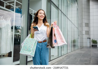 Asian Beautiful Woman Holding Shopping Bag And Smartphone On Her Hands Near The Mall Shop Window, Smiling Shopper Young Girl Standing Using Mobile Phone After Shopping On City Street