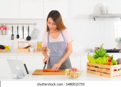Asian Beautiful Woman, Long Hair, Wearing A Pink T-shirt And Striped Apron Standing, Slicing Carrots On The Table At Modern White Kitchen, And Watching Cooking Video Clip Using Tablet.